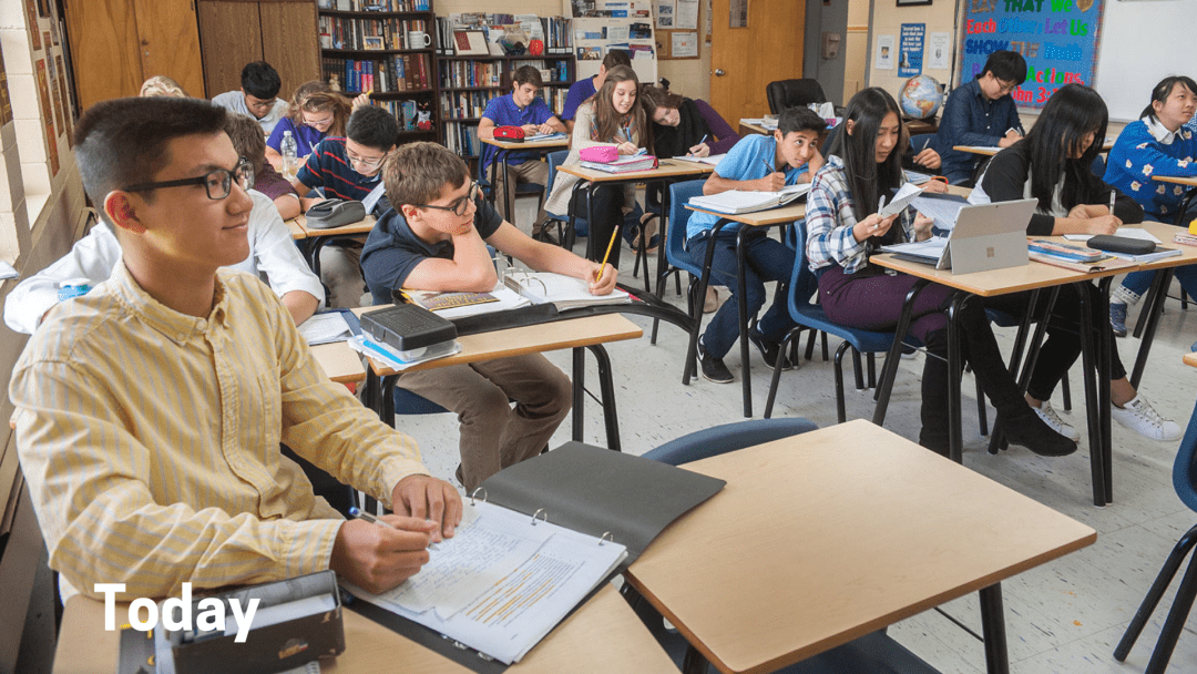 A modern classroom with students sitting in desks in rows. One student has a laptop on her desk.