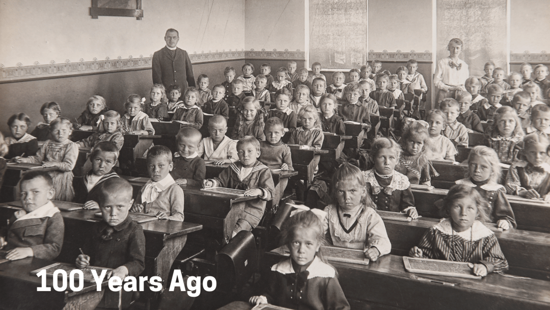 An old black and white photograph of a large classroom crammed with students sitting neatly in rows in long desks facing the front of the room.